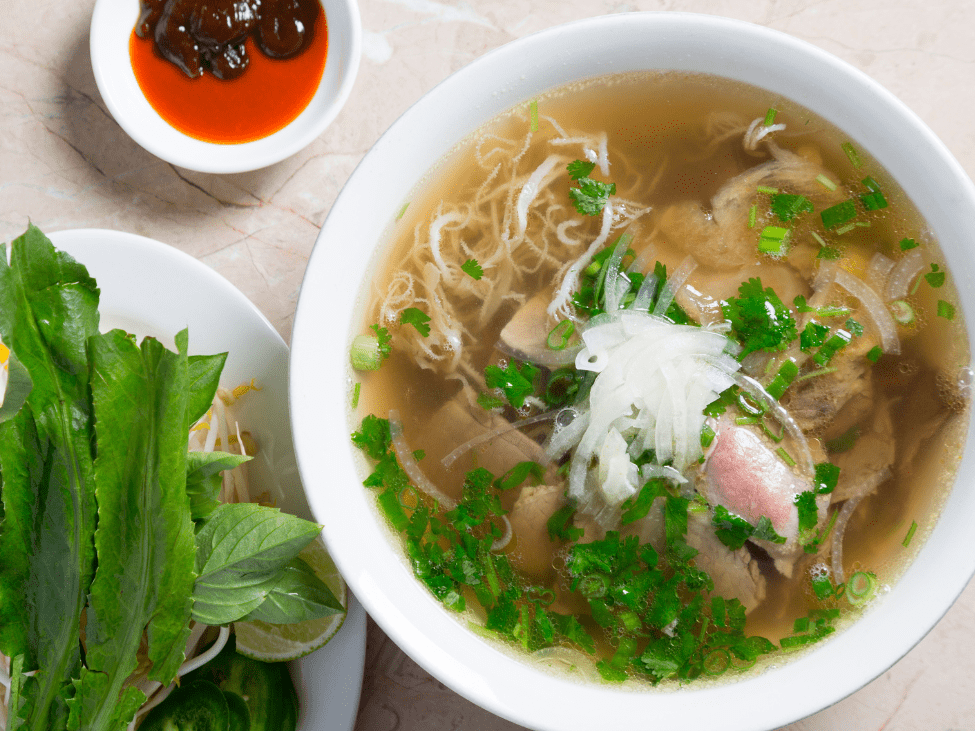 A serene riverside setting in Can Tho, with a steaming bowl of pho enjoyed by the water’s edge, capturing the local culinary experience.