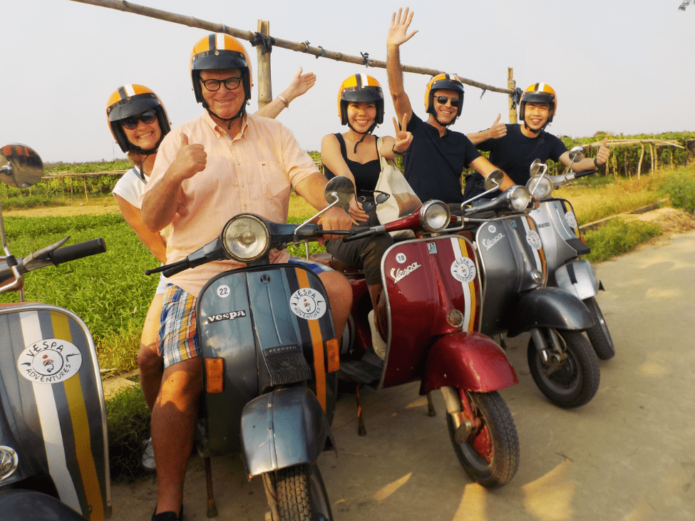 Vespa riders taking a break in a picturesque village in Hoi An, enjoying local snacks and views of rice fields.