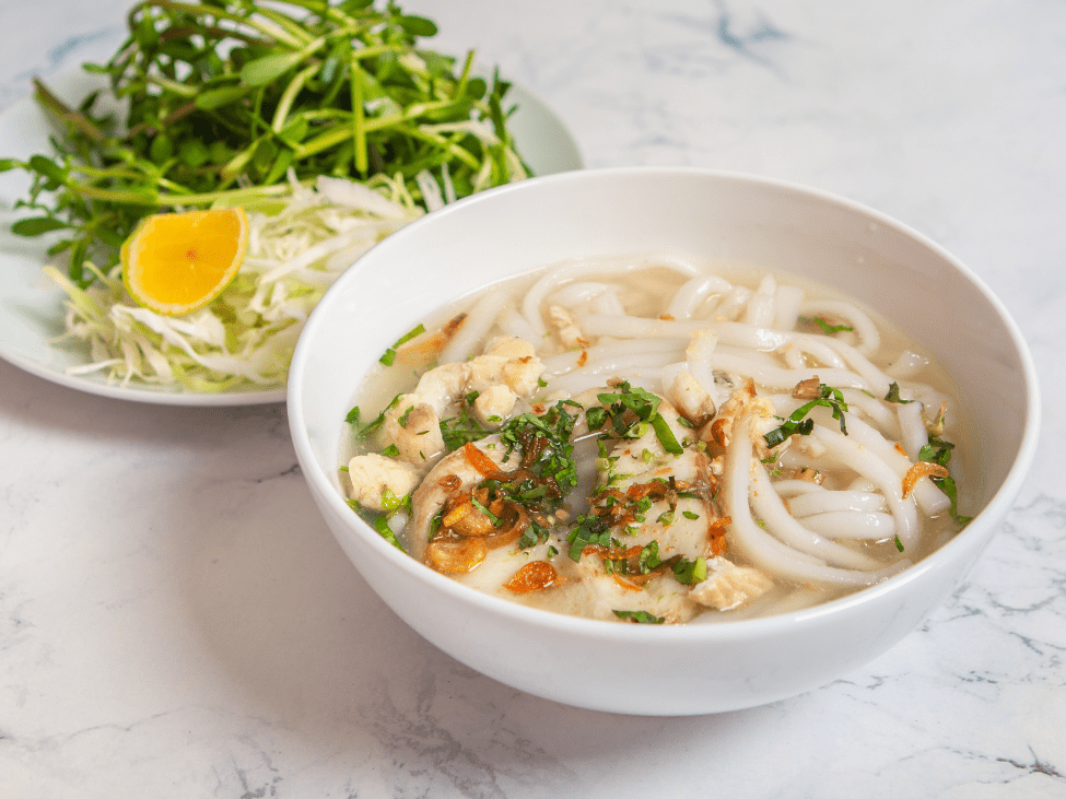 A bowl of banh canh with shrimp and thick tapioca noodles, garnished with herbs and chili.