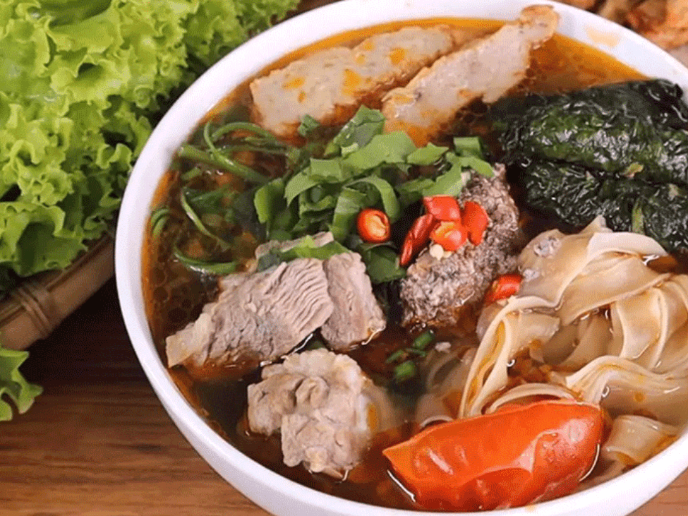 A bowl of banh da cua with brown rice noodles, crab, and beef, topped with fresh herbs.