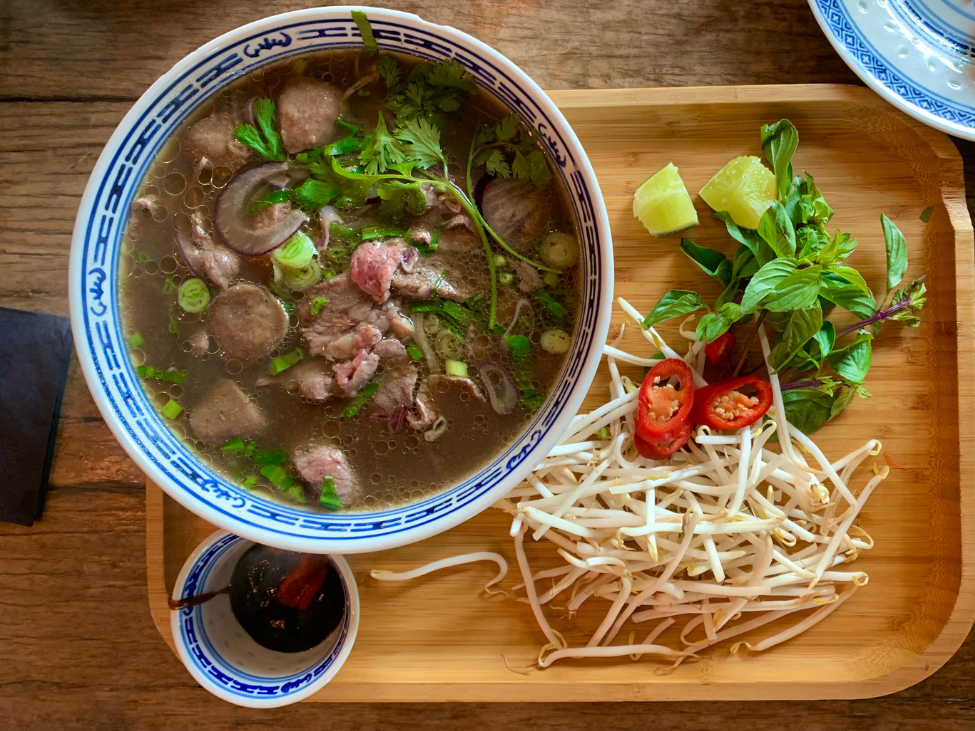 A bowl of beef pho with sliced beef, rice noodles, and fresh herbs, accompanied by lime wedges and chili sauce.