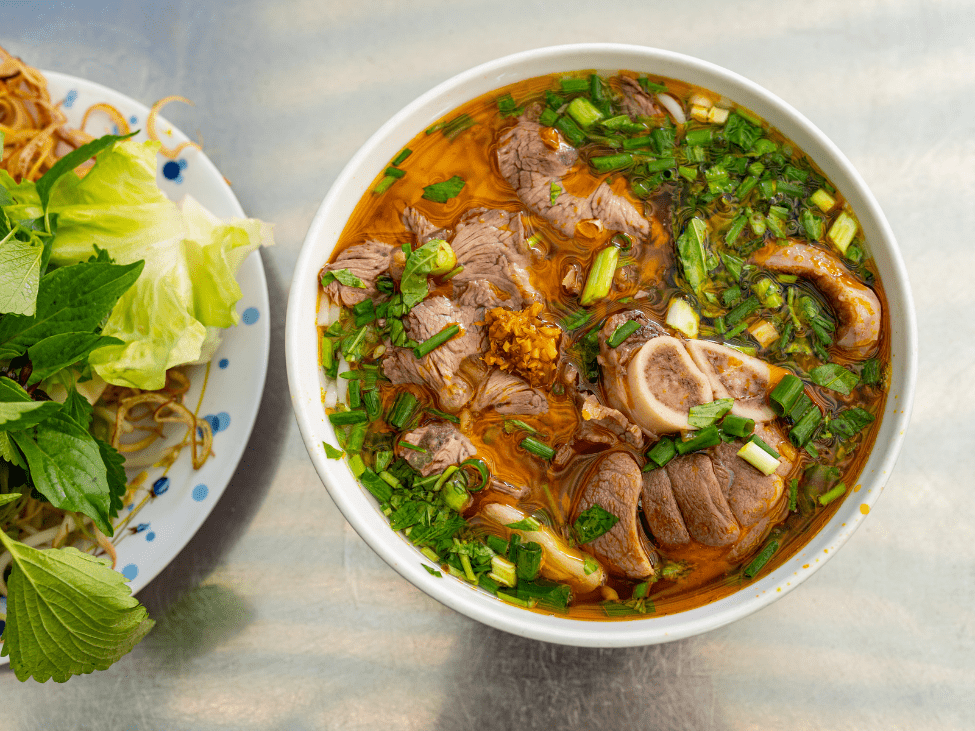 A steaming bowl of bun bo hue with slices of beef and pork, garnished with fresh herbs and a side of lime.