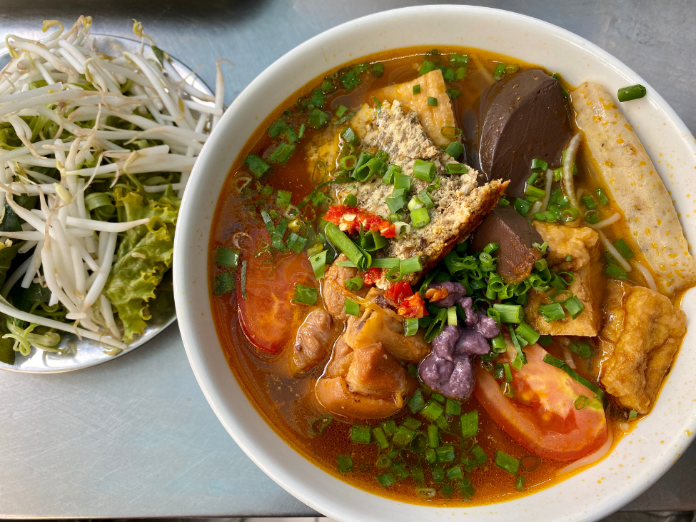 A bowl of bun rieu with crab paste, tofu, and tomatoes, topped with herbs and lime.