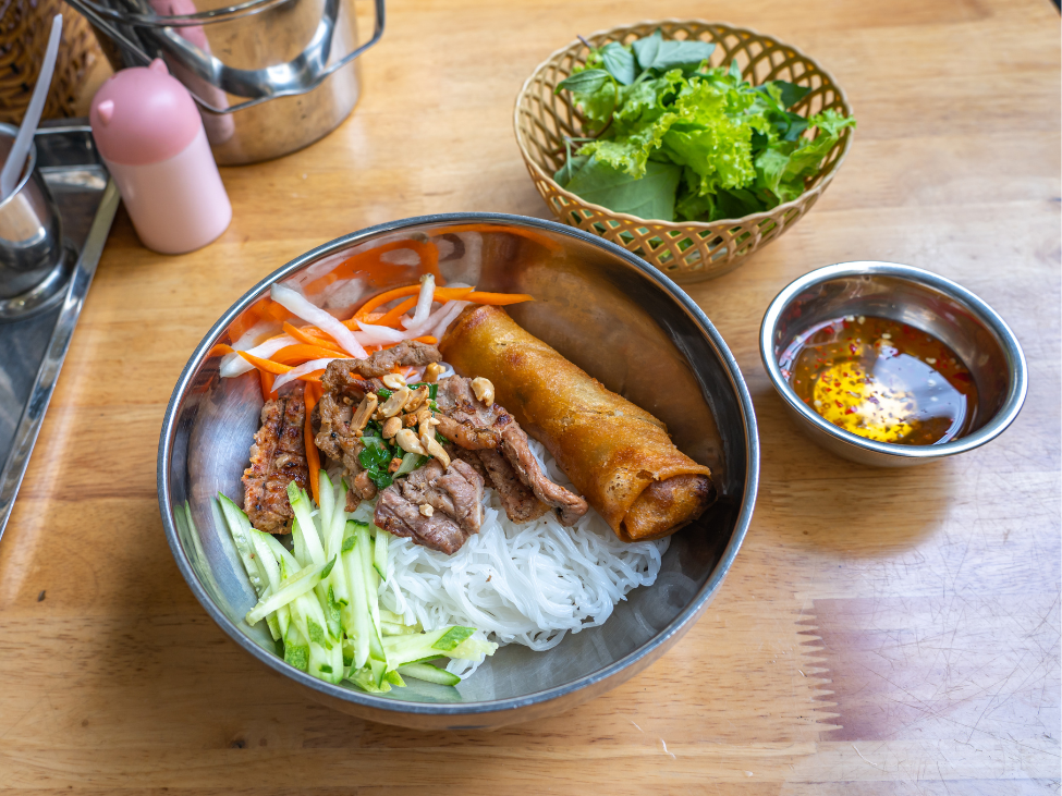 A bowl of bun thit nuong with grilled pork, vermicelli noodles, and fresh herbs, served with a side of fish sauce.