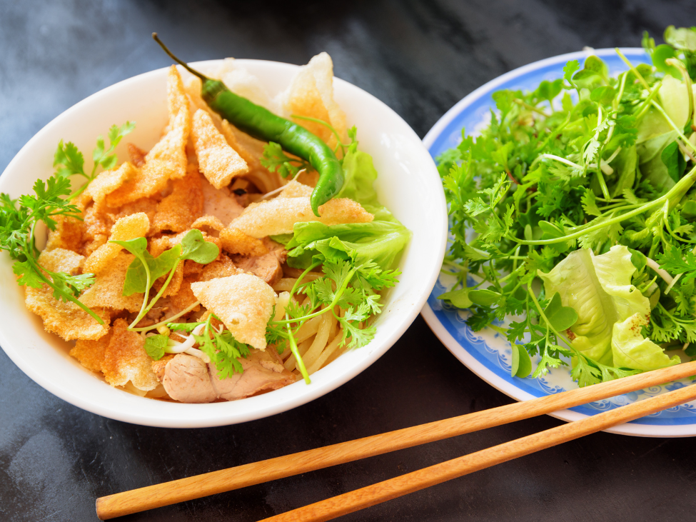 A bowl of cao lau with barbecue pork, fresh greens, and crispy croutons, set in a rustic setting.