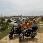 Family crossing a river on a Vespa in Hoi An countryside, surrounded by lush greenery and rice paddies.