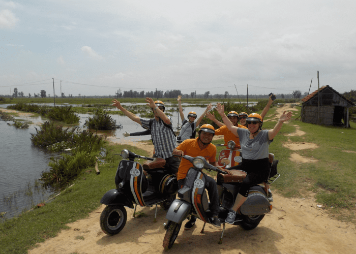 Family crossing a river on a Vespa in Hoi An countryside, surrounded by lush greenery and rice paddies.