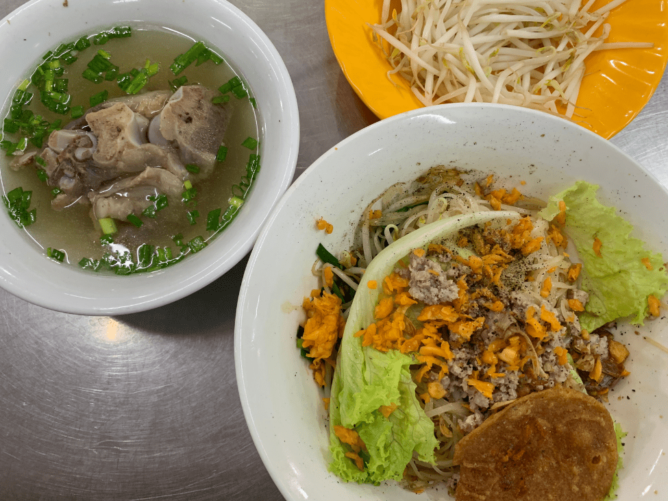 A bowl of hu tieu with thin rice noodles, pork, and fresh herbs, accompanied by a side of dipping sauce.