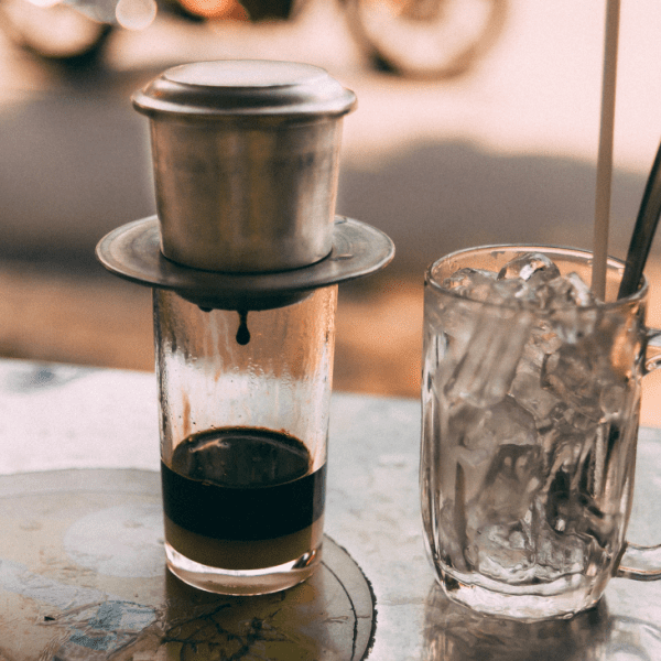 A glass of iced black coffee with a metal straw, placed on a wooden table with coffee beans scattered around.