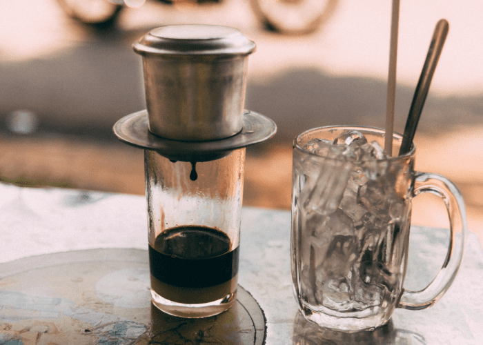 A glass of iced black coffee with a metal straw, placed on a wooden table with coffee beans scattered around.