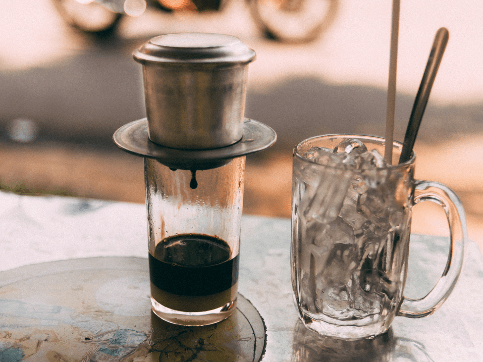 A glass of iced black coffee with a metal straw, placed on a wooden table with coffee beans scattered around.