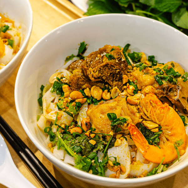 A bowl of mi quang with shrimp, pork, and wide rice noodles, topped with fresh herbs and peanuts.