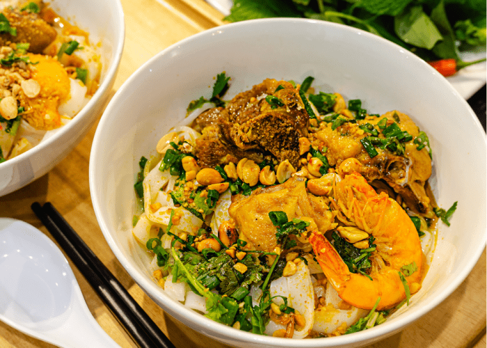 A bowl of mi quang with shrimp, pork, and wide rice noodles, topped with fresh herbs and peanuts.