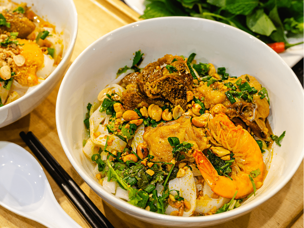 A bowl of mi quang with shrimp, pork, and wide rice noodles, topped with fresh herbs and peanuts.