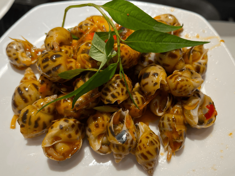 A plate of stir-fried snails with tamarind at Ốc Loan, garnished with fresh herbs and grilled prawns on the side.