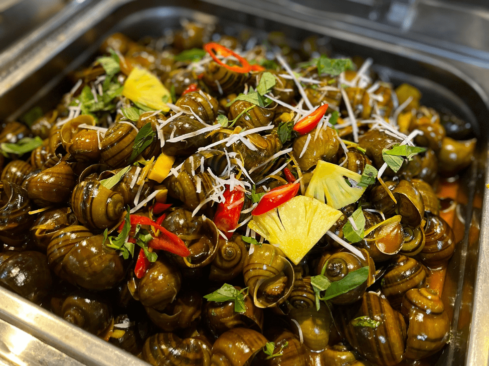 A bowl of snails in tamarind sauce, garnished with peanuts and chili, alongside grilled prawns and squid.