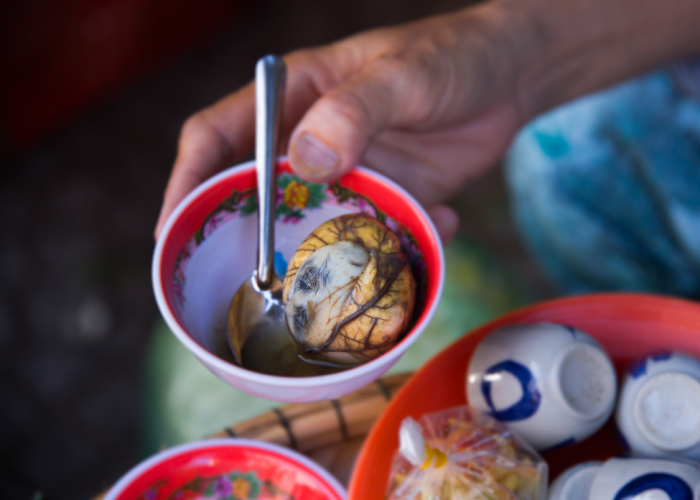 Close-up image of a Vietnamese balut egg with a partially developed embryo visible, served with salt, pepper, and fresh herbs.