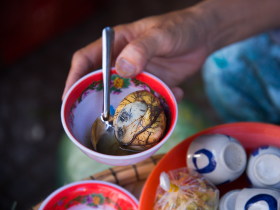 Close-up image of a Vietnamese balut egg with a partially developed embryo visible, served with salt, pepper, and fresh herbs.