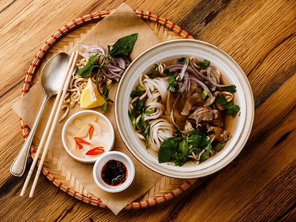 A steaming bowl of Vietnamese pho noodle soup with fresh herbs and lime wedges.