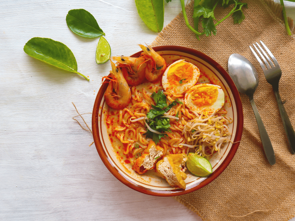 A steaming bowl of laksa noodle soup garnished with bean sprouts and fresh herbs.