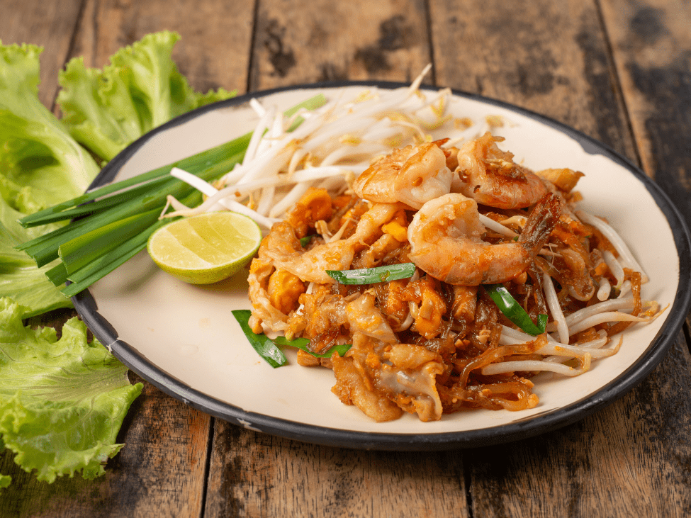 A plate of pad Thai served with shrimp, fresh lime, and crushed peanuts in a bustling Bangkok market.